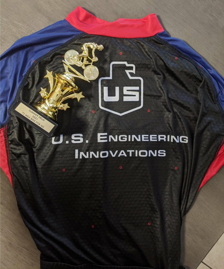 A black and red cycling jersey with U.S. Engineering Innovations text and logo is laid flat, with a gold cycling trophy placed on top.