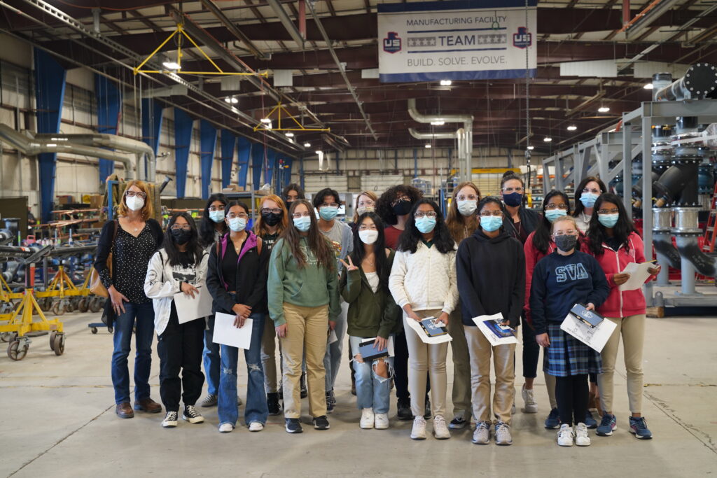 A group of students and an adult stand in a manufacturing facility, all wearing face masks. They hold papers and pose for a group photo in front of industrial equipment and machinery.