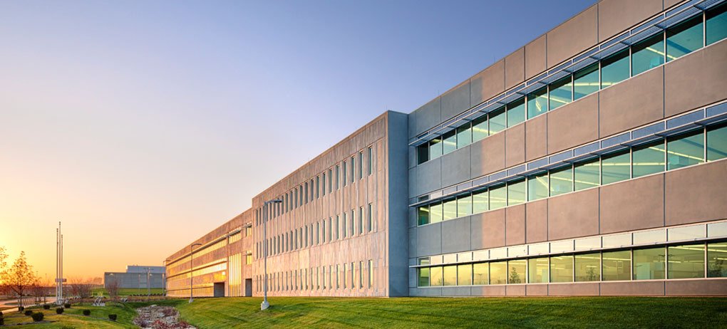 A modern, multi-story office building with large windows and a concrete exterior sits on a well-maintained grassy lawn at sunset. The sky is clear with a warm glow from the setting sun.