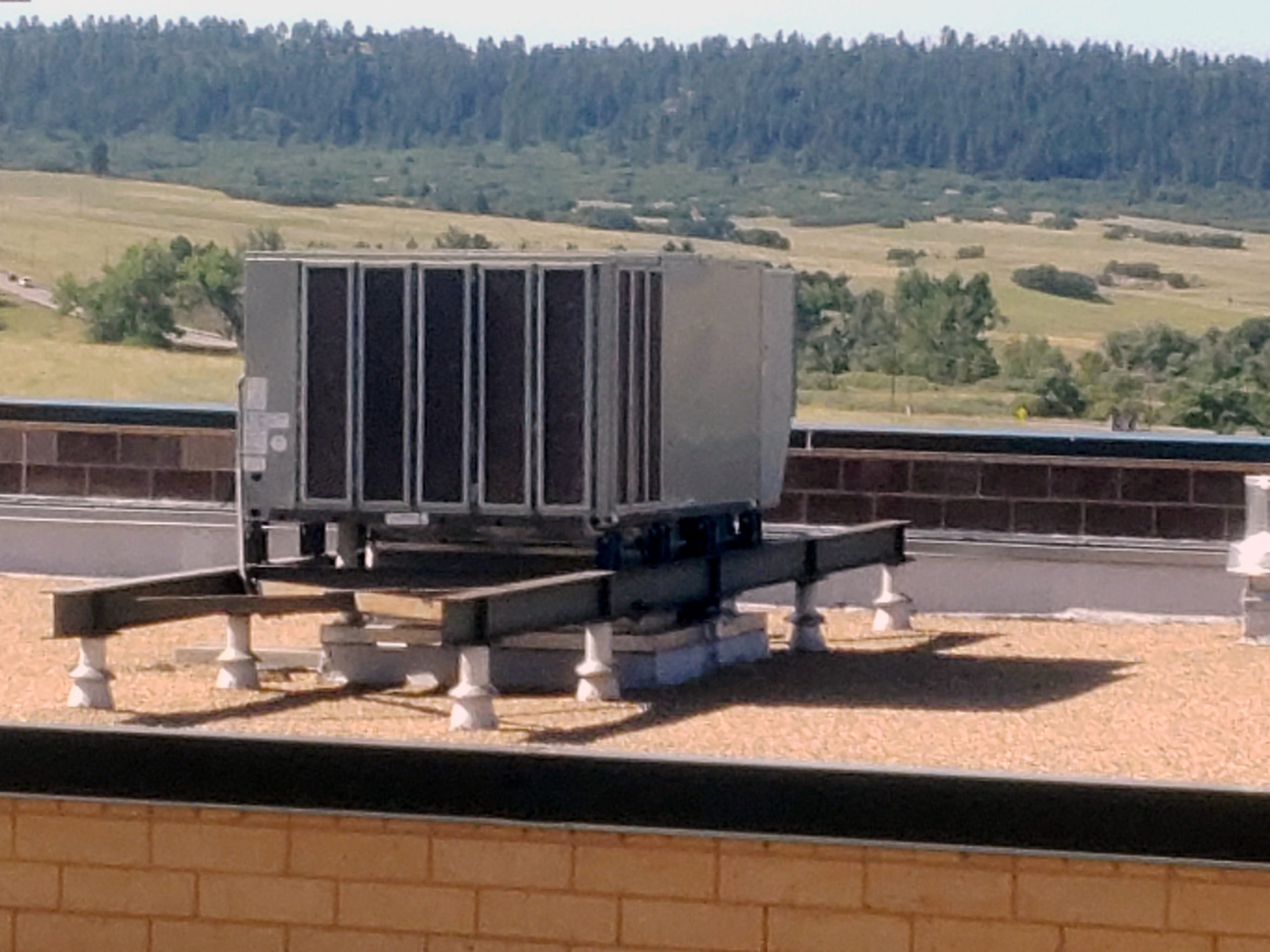 A large HVAC unit is installed on a rooftop, elevated on a metal frame with support blocks. In the background, trees and grassy hills are visible under a clear sky.
