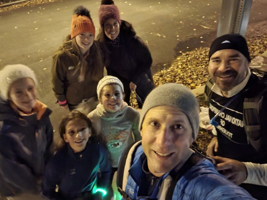 A group of seven people dressed in warm clothes pose for a selfie outdoors at night. They are standing on a street near fallen autumn leaves, and some are wearing hats and gloves. Everyone is smiling at the camera.