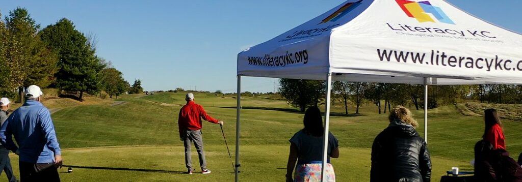A group of people stands on a golf course near a white canopy tent with the Literacy KC logo and website. One person is golfing while others watch under a clear, sunny sky.