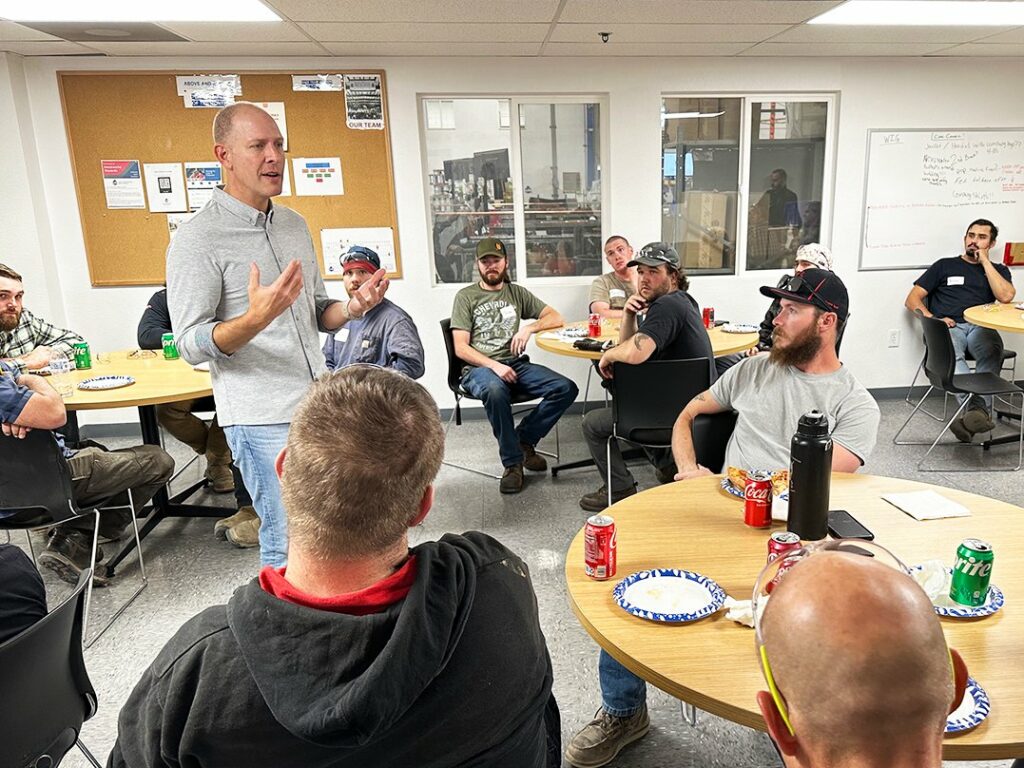 A man stands and speaks to a group of people seated at round tables in a break room. Some attendees have drinks and food in front of them, and bulletin boards and windows are visible in the background.
