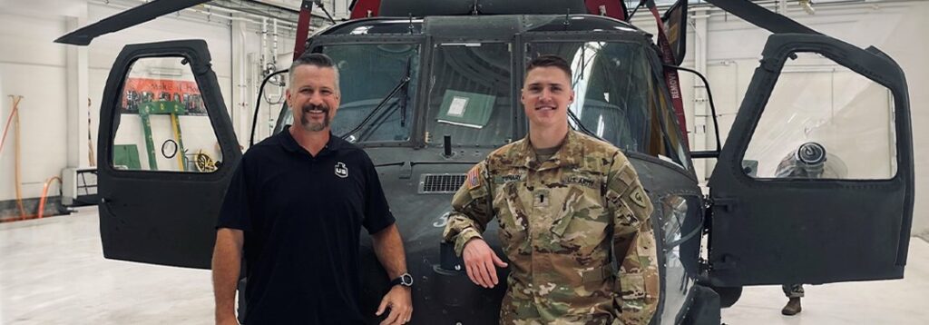 Two people stand in front of a military helicopter inside a hangar. One wears a black polo shirt and the other is in camouflage military uniform. The helicopter’s doors are open, and equipment is visible in the background.