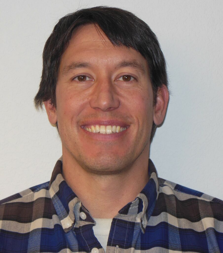 A man with straight dark hair is smiling at the camera. He is wearing a blue and white plaid shirt. The background is plain and light-colored.
