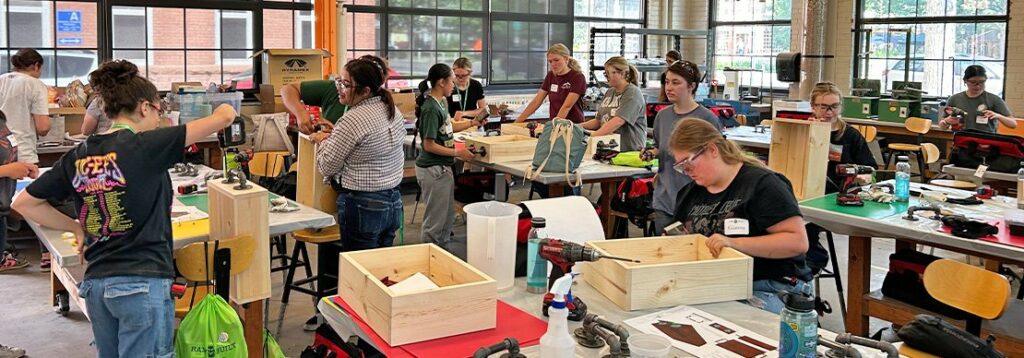 A group of people work on woodworking projects at tables in a bright, open workshop. Various tools and unfinished wooden boxes are on the tables. Large windows let in natural light.