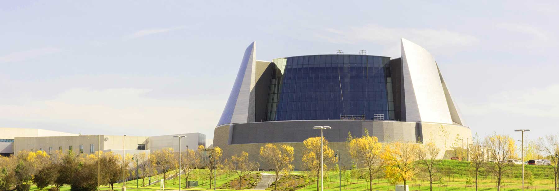 A modern building with a curved glass facade and large silver panels on each side, surrounded by green grass and trees with yellow leaves under a clear sky.