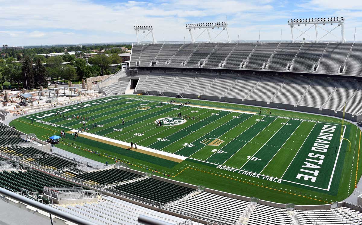 A wide view of Sonny Lubick Field at Colorado State Universitys football stadium, showing green turf with yard lines, stadium seats, and a few people on the field under a partly cloudy sky.