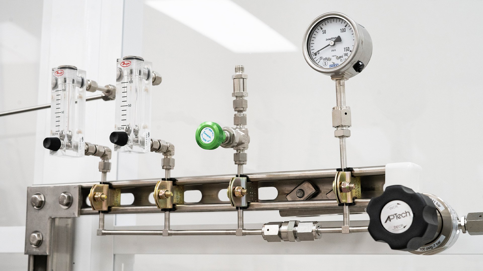 A gas manifold system with two flow meters, a pressure gauge, control valves, and connecting pipes mounted on a metal bracket in a laboratory setting.