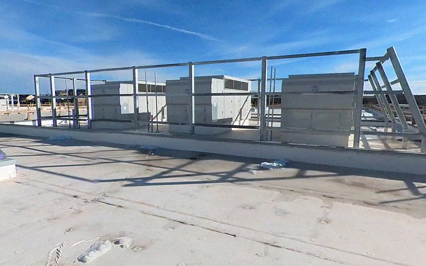 A flat rooftop with HVAC units and metal framing structures under a clear blue sky. Shadows from the structures are cast on the white roof surface. Some construction materials are scattered around.