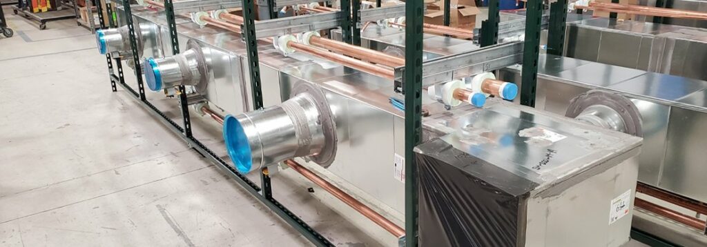 Rows of large metal HVAC duct units with attached pipes and blue plastic covers on the vents are arranged in a warehouse or workshop setting.