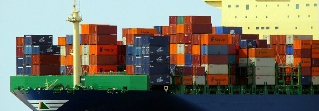 A large cargo ship loaded with colorful shipping containers stacked in rows is shown at sea. The ships hull is dark blue with a green deck area and a yellow superstructure is partially visible at the top right.