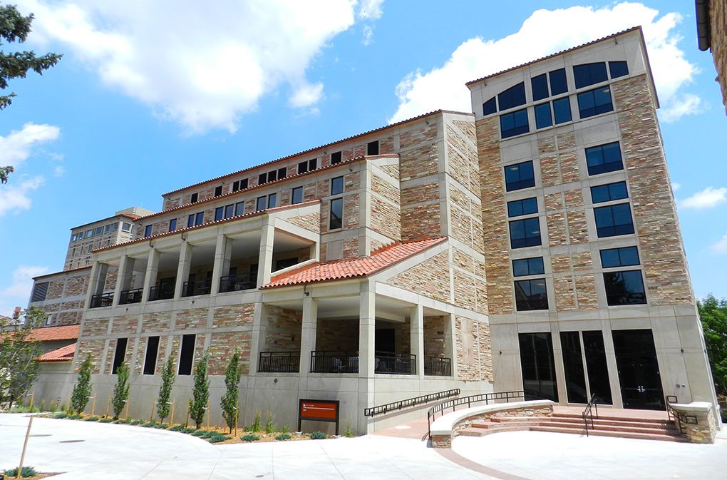 A five-story building with tan stone walls, large windows, and red-tiled roofs stands under a blue sky with scattered clouds. The structure has multiple levels, columns, and a small sign near the entrance.