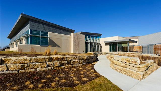 A modern building with large windows and geometric shapes, surrounded by landscaped stone walls, brown mulch, and a curved concrete walkway under a clear blue sky.