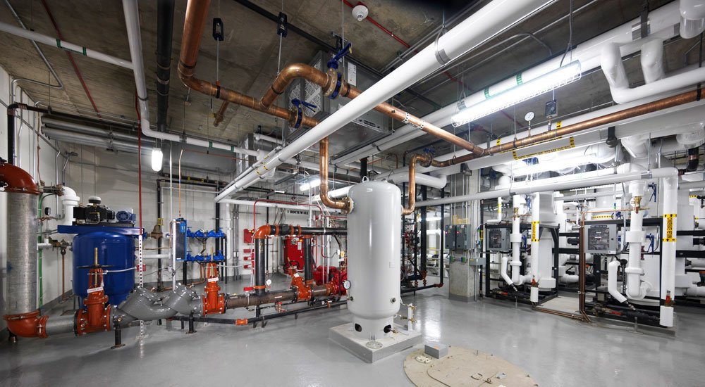 A mechanical room with exposed pipes, tanks, valves, and industrial equipment. The floor is clean and gray, and the ceiling shows structural concrete and additional piping. The lighting is bright and industrial.