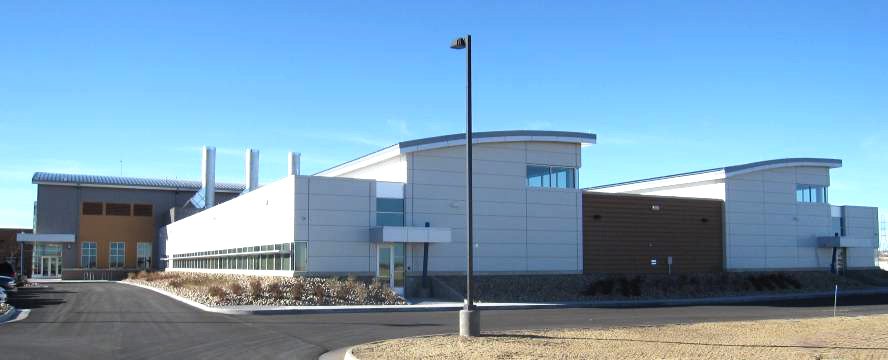 A modern, single-story building with a flat roof and white exterior walls, featuring large windows and multiple chimneys, sits under a clear blue sky with minimal landscaping and a paved road in front.