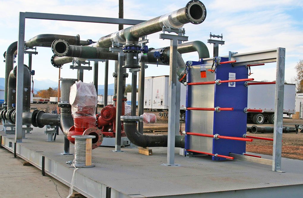 An industrial skid-mounted heat exchanger system with black pipes, a red pump, and a blue plate heat exchanger, installed outdoors on a metal platform. Several trucks are visible in the background.