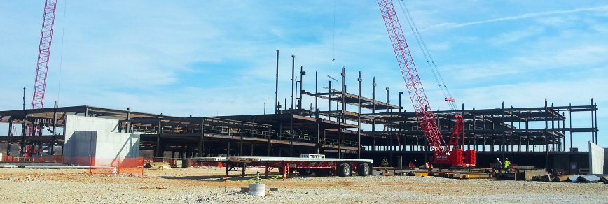 A large building under construction with exposed steel framework, a red crane, construction materials, a trailer, and a few workers visible on a dirt-covered work site under a blue sky.