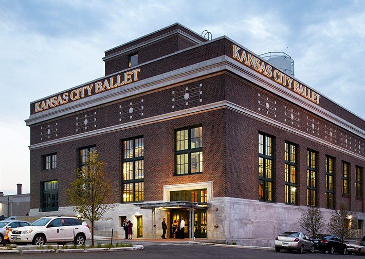 A brick building with large windows and Kansas City Ballet signage on two sides. Several cars are parked in front, and a few people are entering through the main entrance. The sky is mostly cloudy.