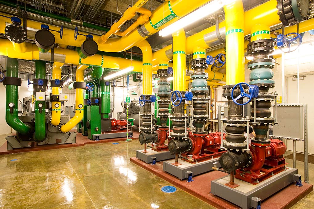 A brightly lit mechanical room featuring multiple large yellow and green pipes, red pumps, black valves, and blue handwheels installed on a polished concrete floor. The space is clean and organized.