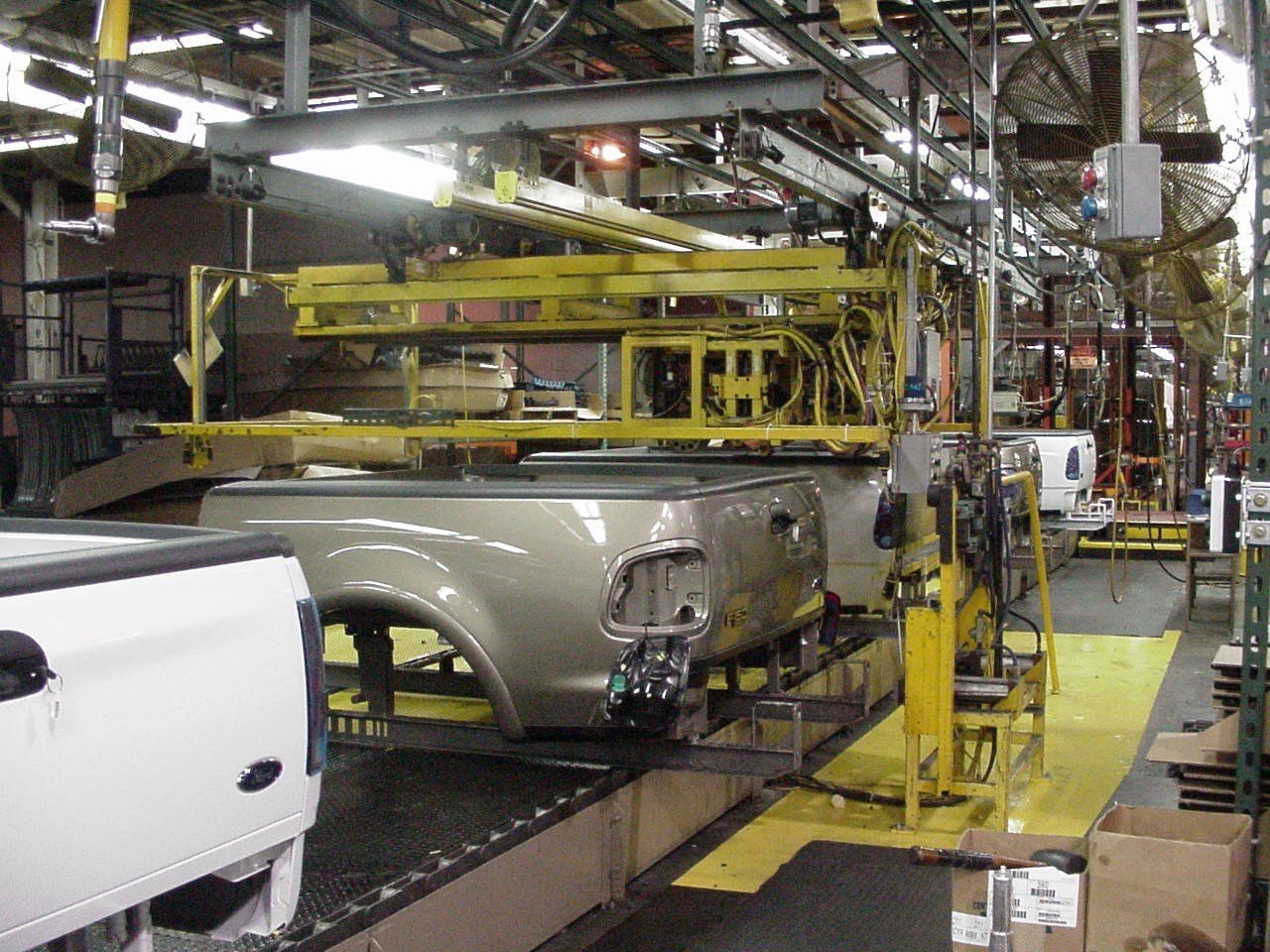 An automotive assembly line with truck beds being transported by overhead machinery inside a factory. The environment is industrial, with metal structures, yellow equipment, and various parts in progress.