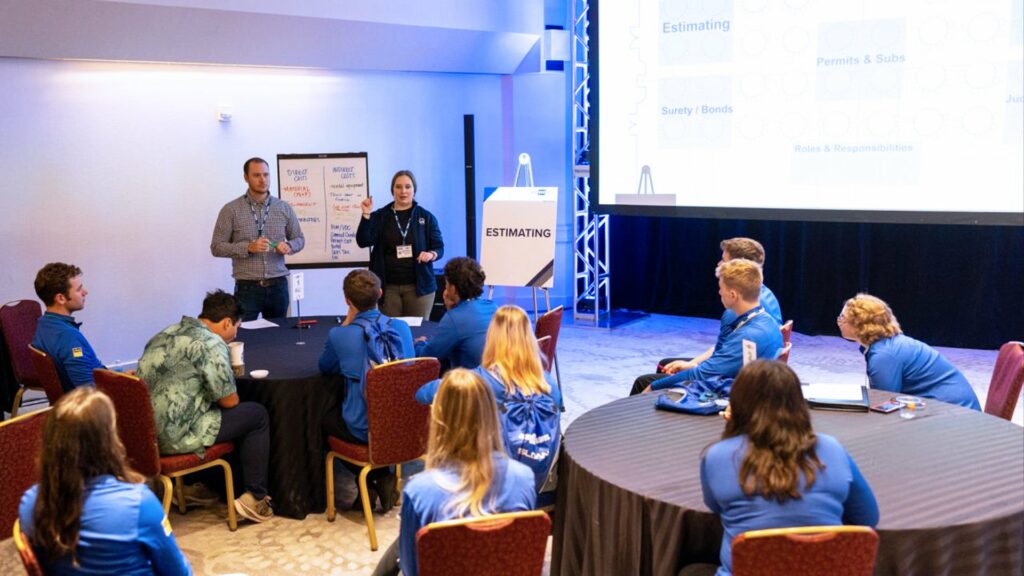 A group of people in blue shirts sit at round tables, facing two presenters standing at the front of a conference room. A presentation is displayed on a screen, and a flip chart labeled “Estimating” is visible.