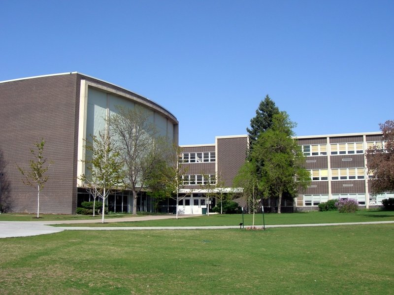 A three-story brick school building with large windows and an entrance at the center, surrounded by a grassy lawn and several trees, under a clear blue sky.