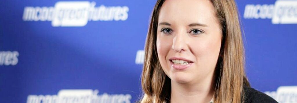 A woman with straight light brown hair is speaking in front of a blue backdrop with white text that reads mcon great futures in a repeated pattern. She is wearing a dark blazer and a white top.