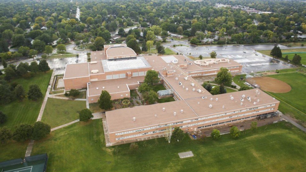 Aerial view of a large brick school building surrounded by grass fields, trees, a baseball diamond, and parking areas, with residential houses and roads in the background.