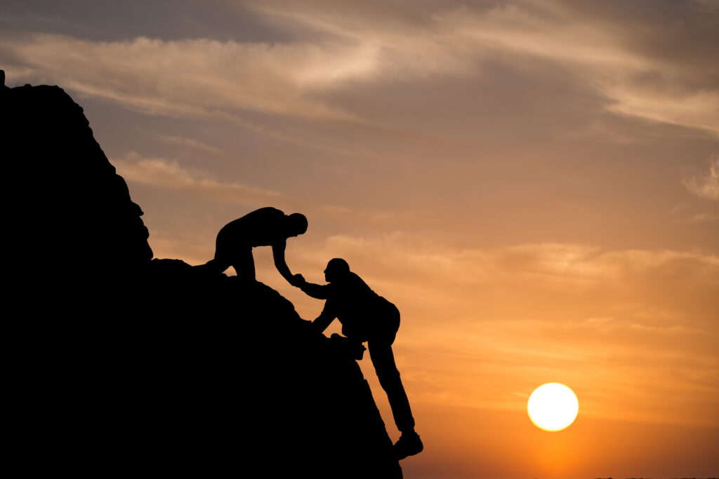 Two people are silhouetted against a sunset sky while climbing a rocky slope; one person extends a hand to help the other up the incline. The sun is low on the horizon.