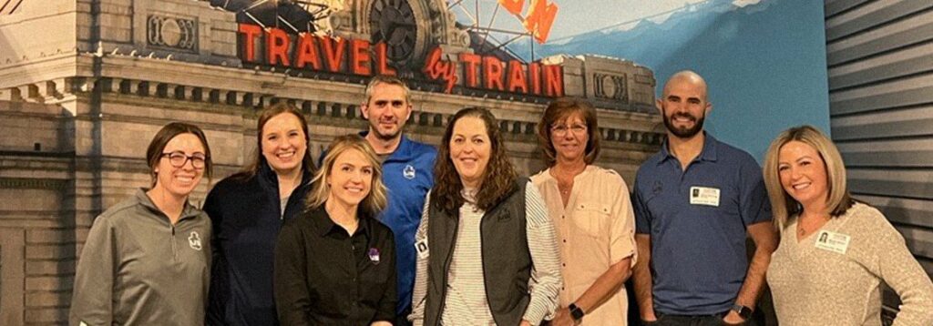 Eight people stand indoors in front of a mural of a train station with the words “Travel by Train.” Most are smiling and wearing business casual attire. Some have name badges. The setting appears to be a professional gathering.
