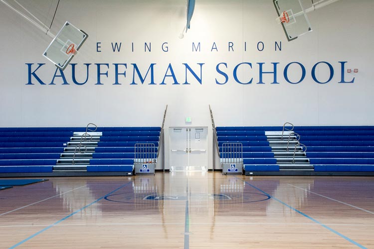 View of a gymnasium with blue bleachers and a polished wooden floor. Ewing Marion Kauffman School is written on the wall above double doors at the center of the bleachers. Two basketball hoops are visible.