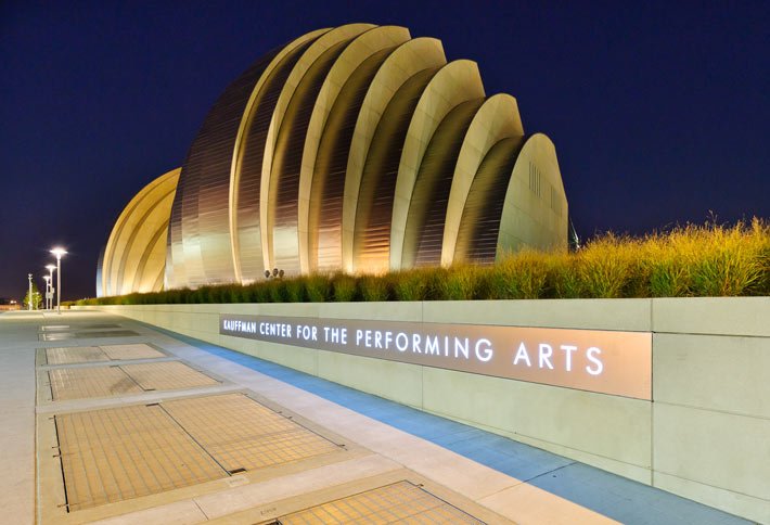 The Kauffman Center for the Performing Arts at night, featuring its distinctive curved, shell-like architecture and illuminated signage in front of landscaped grass.