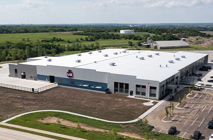 A large, white industrial building with a flat roof and multiple rooftop vents is surrounded by parking lots and green fields under a partly cloudy sky. There is a company logo on the front of the building.