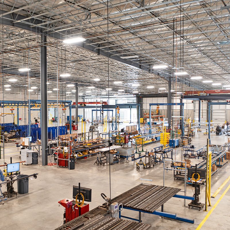 Large, well-lit factory floor with high ceilings, metal support beams, conveyor belts, machinery, workstations, and various industrial equipment organized throughout the open space.