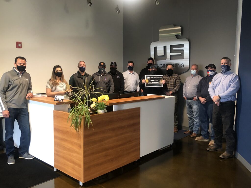 A group of twelve people wearing masks stands in an office lobby behind a reception desk with yellow flowers. A large metallic US Engineering sign is mounted on the wall behind them.