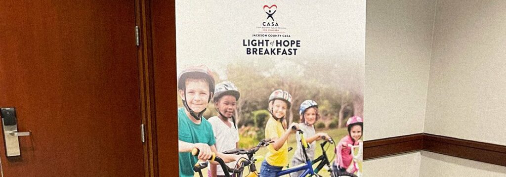 A standing poster in a hallway displays the CASA organization logo and the words Light of Hope Breakfast above a photo of five children outdoors, lined up wearing helmets and holding bicycles.
