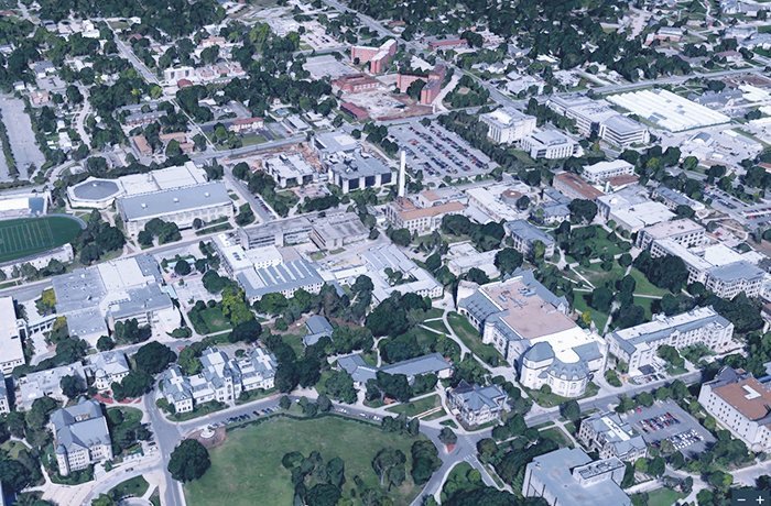 Aerial view of a university campus showing academic buildings, roads, green spaces, and surrounding residential areas. Trees are scattered throughout the campus, and several parking lots are visible.