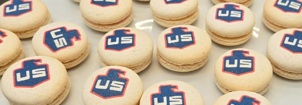 A group of light-colored macarons arranged on a tray, each featuring the letters US in white on a blue and red shield-shaped background printed on top.