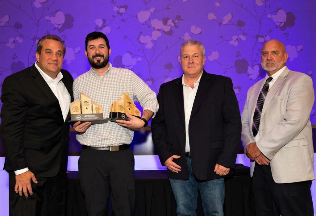 Four men pose for a photo at an indoor event. The man second from left holds several wooden award plaques. All are dressed in suits or business attire, standing in front of a purple floral-patterned wall.