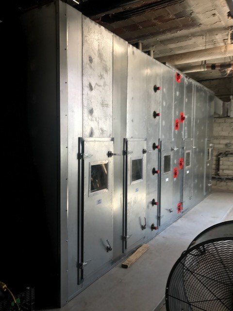 A large industrial metal ventilation system with access panels and small windows is installed along a wall in a utility room. Red valve handles and visible pipes are attached to the system.