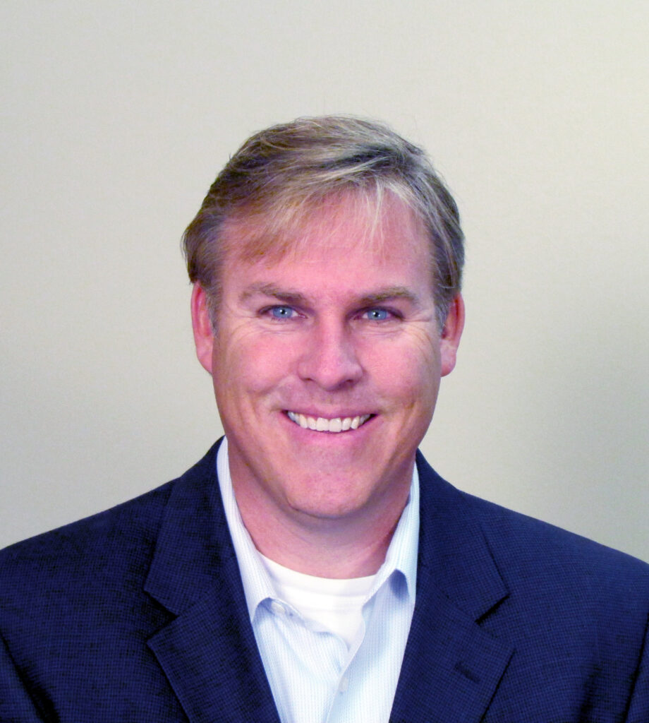 A man with light brown hair wearing a dark jacket over a light collared shirt is smiling at the camera against a plain, light-colored background.