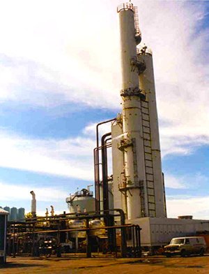 A tall, white industrial distillation tower with pipes and ladders stands at a refinery site under a partly cloudy sky. Smaller structures and a white van are visible in the foreground.