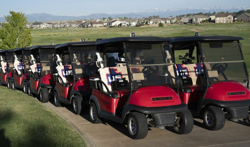 A row of red golf carts is parked along a sidewalk on a golf course, with houses and distant mountains visible in the background under a clear sky.