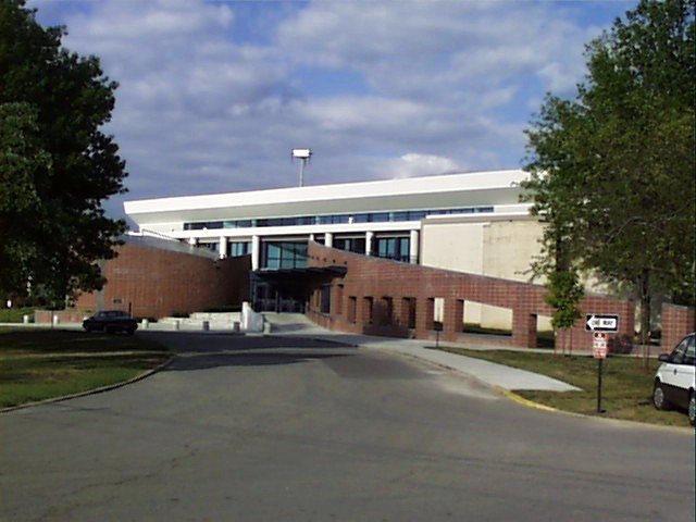 A large, modern building with a brick ramp entrance and glass doors is surrounded by trees and a paved road. The sky is partly cloudy, and a few parked cars are visible near the entrance.