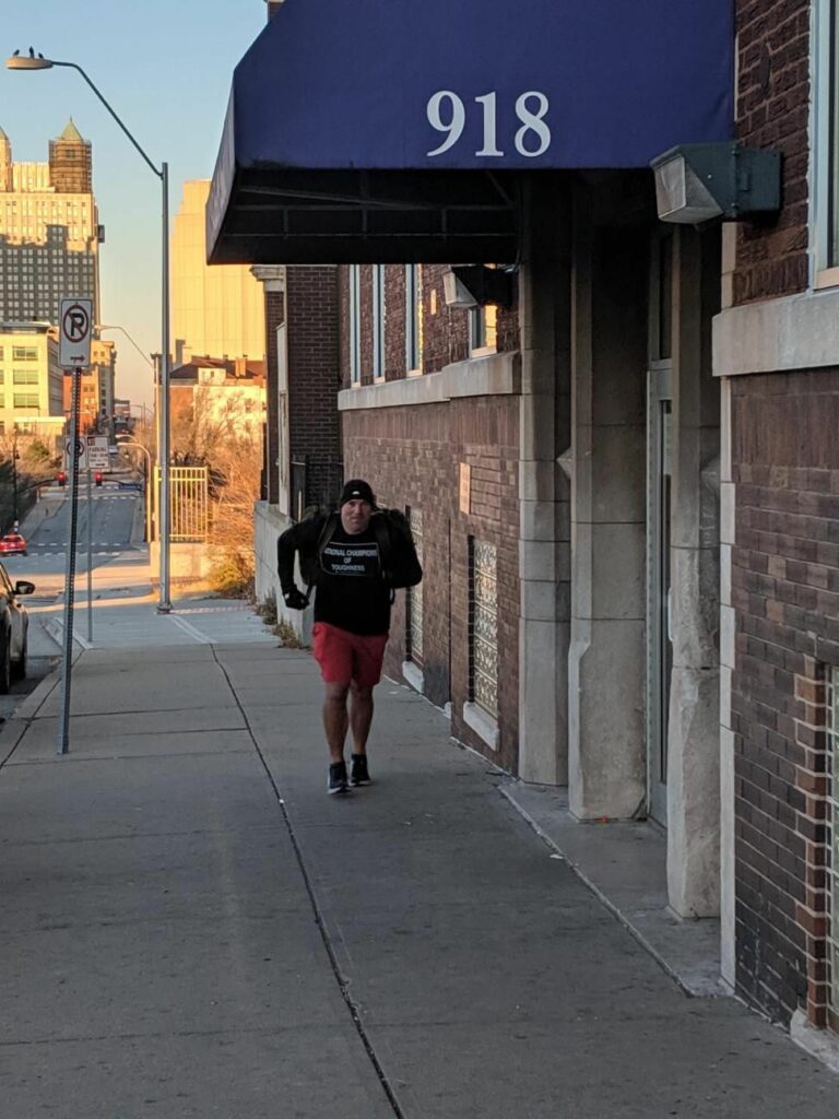 A person wearing a black shirt, red shorts, and a black beanie walks on a sidewalk next to a brick building with the number 918 above the entrance. City buildings and a street are visible in the background.