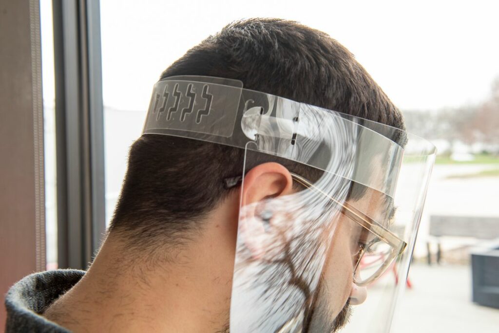 A person wearing glasses and a transparent face shield is standing indoors near a window, facing sideways. The face shield covers their entire face and is secured with an adjustable headband.