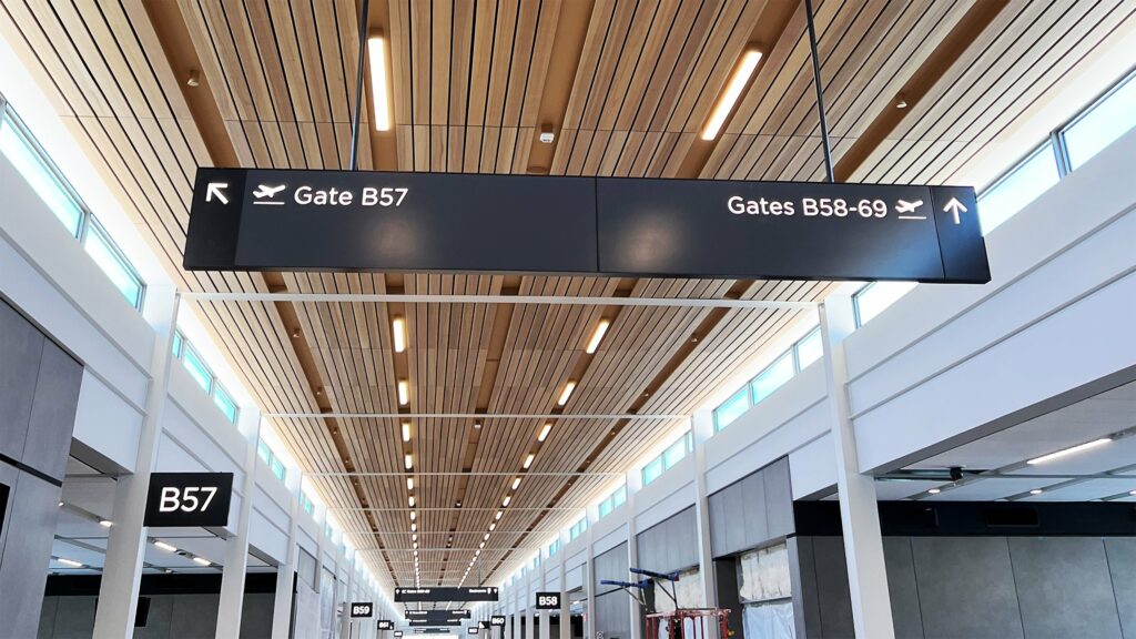 Overhead airport signs point left to Gate B57 and straight ahead to Gates B58-69. The signs hang from a ceiling with wooden slats. The terminal appears well-lit and modern. Gate B57 sign is also visible on the left wall.