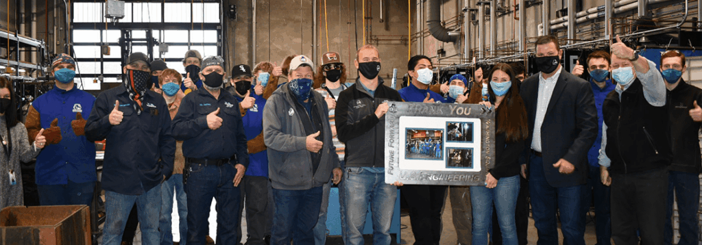 A group of people wearing masks stand indoors, some giving thumbs up. One person in the center holds a framed display with photos and the words “THANK YOU” on it. Industrial equipment is visible in the background.