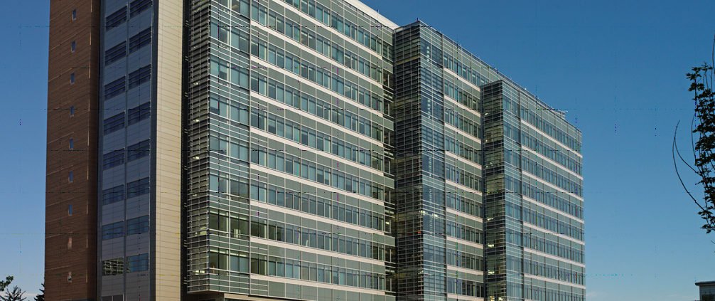A modern, multi-story office building with glass and metal exterior panels, horizontal window bands, and a vertical section on the left; clear blue sky in the background.